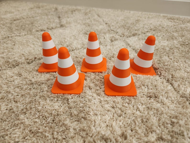 May include: Five orange and white striped traffic cones. Each cone has a square orange base and a conical top with three white stripes. The cones are arranged on a beige carpet.