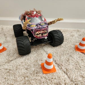May include: A toy monster truck with a zombie-themed design, featuring a detailed zombie face and arms. The truck has large black tires and is surrounded by orange and white traffic cones, creating a playful scene.