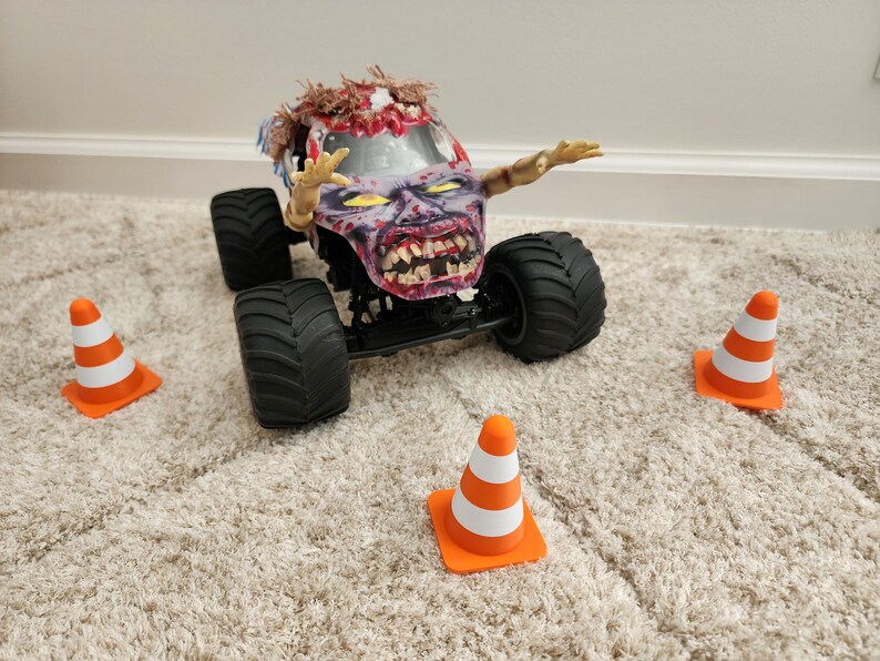 May include: A toy monster truck with a zombie-themed design, featuring a detailed zombie face and arms. The truck has large black tires and is surrounded by orange and white traffic cones, creating a playful scene.