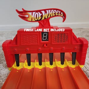 May include: A red Hot Wheels toy race track finish line structure with six orange lanes, a digital display showing the number eight, and a prominent Hot Wheels logo at the top. The text FINISH LANE NOT INCLUDED is printed in white on the front.
