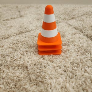May include: A small orange and white striped traffic cone. The cone has a rounded top and is sitting on a stack of three orange squares. The cone is on a beige, textured surface.