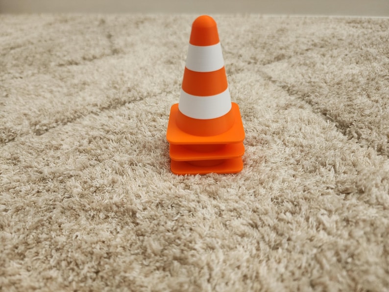 May include: A small orange and white striped traffic cone. The cone has a rounded top and is sitting on a stack of three orange squares. The cone is on a beige, textured surface.
