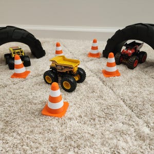 May include: A toy construction site scene with three monster trucks, orange and white traffic cones, and two black tire arches. The trucks are yellow, red, and black. The scene is set on a beige carpet.