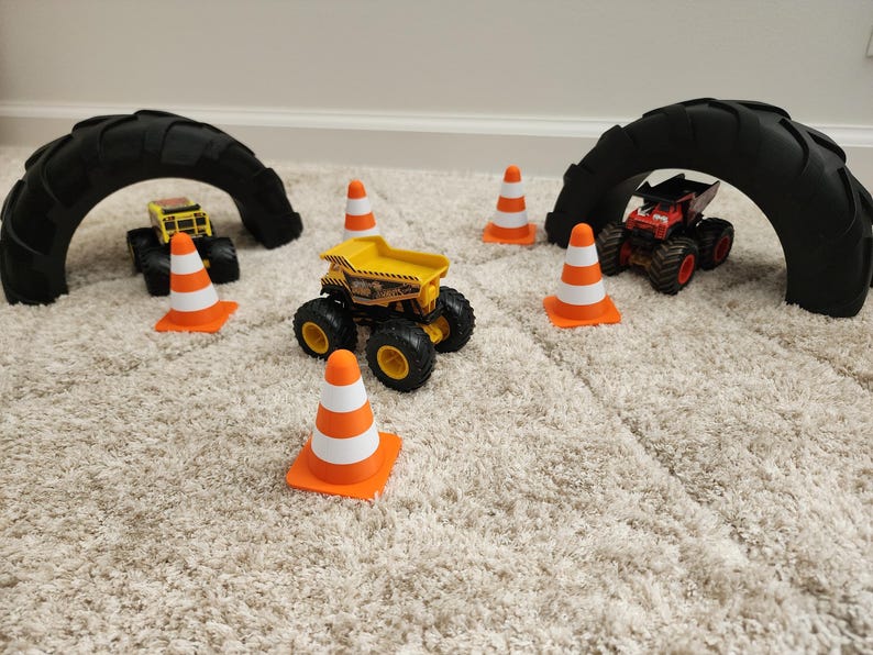 May include: A toy construction site scene with three monster trucks, orange and white traffic cones, and two black tire arches. The trucks are yellow, red, and black. The scene is set on a beige carpet.