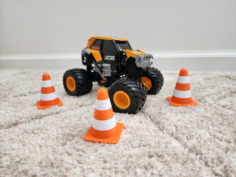 May include: A black and yellow JCB monster truck toy with large black tires and orange rims. Three orange and white traffic cones are placed around the truck on a beige carpet.