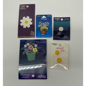 Puede incluir: Una colección de apliques termoadhesivos de la marca Wrights con diseños florales. Los apliques incluyen margaritas y arreglos florales en cestas y macetas. El embalaje es de varios colores, incluyendo morado, azul y blanco.