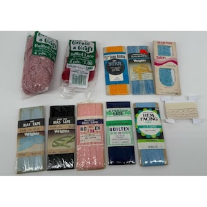 Könnte beinhalten: Eine Sammlung von Nähutensilien, darunter Rüschenspitze, Schrägband, Nahtband und Stretchspitze. Die Artikel sind in verschiedenen Farben verpackt, wie Rosa, Blau, Schwarz und Creme. Die Verpackungen haben Textetiketten.