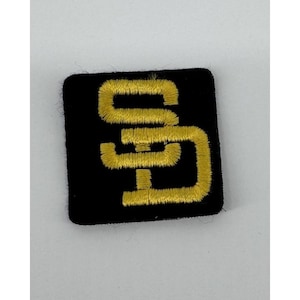 May include: A square black patch featuring a gold embroidered SD logo. The logo is a stylized interlocking design, with the letters "S" and "D" intertwined. The patch is likely for clothing or accessories.