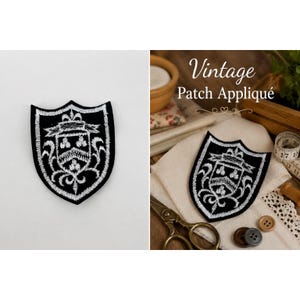 Pode incluir: Um emblema preto em forma de escudo com detalhes heráldicos bordados em branco, incluindo um chevron central e motivos florais. O design inclui uma faixa no topo e volutas complexas, apresentado como uma aplicação têxtil plana.