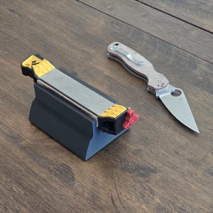 Può includere: Un affilacoltelli nero e giallo con una pietra per affilare grigia e una manopola di regolazione rossa. Un coltello pieghevole argento e beige con una clip da tasca è accanto all'affilacoltelli. La lama del coltello è parzialmente visibile.