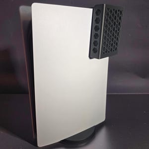 Op de afbeelding: Een witte gameconsole die verticaal op een ronde zwarte voet staat. Aan de rechterbovenkant van de console is een zwart, rechthoekig accessoire bevestigd met een opvallend honingraatpatroon op zowel het voorpaneel als het zijpaneel.