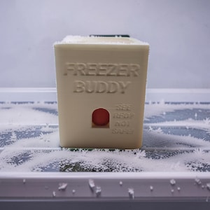 Puede incluir: Un recipiente de plástico beige con la inscripción "FREEZER BUDDY" y un pequeño objeto rojo visible a través de un agujero. El recipiente está dentro de un congelador, rodeado de escarcha.
