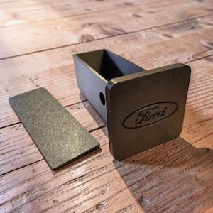 May include: A black matte trailer hitch cover featuring the Ford logo embossed on the front face. The hitch cover has a square-shaped insert and a separate rectangular flat plate with a textured finish, both designed for vehicle towing equipment.