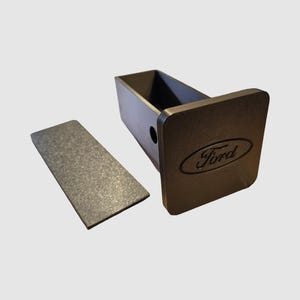 May include: A black metal trailer hitch receiver with the Ford logo embossed on the front. A separate, rectangular metal plate is positioned to the left. The hitch has a square opening and a circular hole.