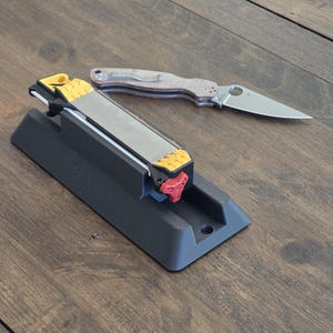 Puede incluir: Un afilador de cuchillos negro con detalles amarillos y rojos, y una navaja plateada con una hoja parcialmente dentada. El afilador tiene una base rectangular y una superficie de afilado metálica. La navaja está abierta, mostrando su hoja.