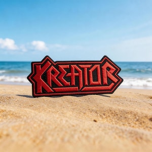 May include: A red and black embroidered patch with the word "KREATOR" in a stylized font. The patch is rectangular with a pointed arrow shape on the right side. The patch is on a sandy beach with the ocean and blue sky in the background.