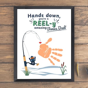Può includere: Una stampa incorniciata con il testo "Hands down, you're a REEL-y amazing Bonus Dad!" presenta un'illustrazione di canna da pesca e un'impronta della mano. L'opera d'arte include un mulinello, un galleggiante e giunchi, il tutto su sfondo bianco.