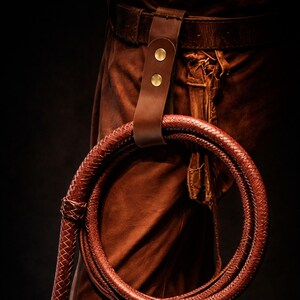 May include: A brown leather whip with a braided design, looped and held by a leather belt loop with two brass rivets. The whip is a reddish-brown color, and the background is dark.