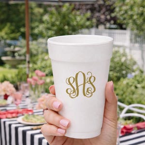 May include: A white disposable cup with a gold monogram design. The cup is held by a person with pink nail polish, with a black and white striped table in the background. An outdoor setting with a striped umbrella is visible.