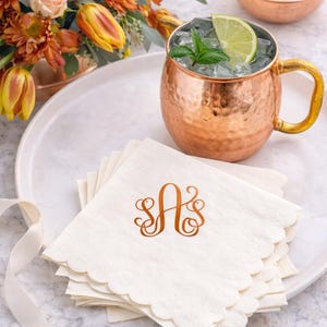 May include: A copper Moscow Mule mug with a gold handle, filled with ice, a lime wedge, and mint. A stack of white scalloped-edge napkins with a copper monogram sits on a white plate. A floral arrangement and sugar cubes complete the scene.