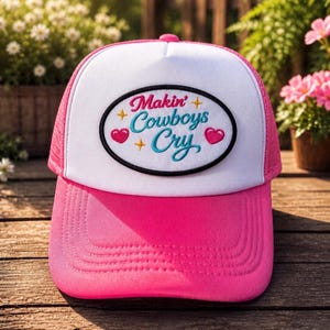 Puede incluir: Gorra de camionero rosa y blanca con el texto bordado "Makin' Cowboys Cry". La gorra tiene una visera rosa y una parte trasera de malla, con un panel frontal blanco. El texto está en una fuente cursiva con acentos de corazón y estrella.