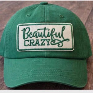 May include: A green baseball cap with a curved brim. The cap features a white rectangular patch with the words "Beautiful Crazy" in a cursive font. The cap is made of a soft fabric.