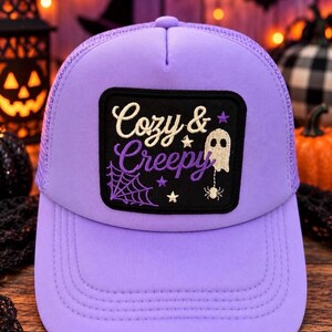 Puede incluir: Una gorra tipo trucker de color morado claro con un parche rectangular negro en la parte frontal. El parche muestra el texto bordado "Cozy & Creepy" en blanco y morado, junto a un fantasma blanco, una araña, una telaraña y estrellas.