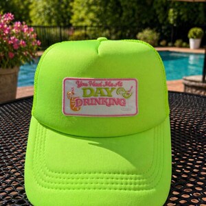 Könnte beinhalten: Leuchtend neongrüne Trucker-Cap mit Mesh-Rückseite und gebogenem Schirm. Auf der Vorderseite befindet sich ein rechteckiger Aufnäher mit dem Text "You Had Me At DAY DRINKING" und Cocktail-Illustrationen. Die Kappe ist für den Freizeitgebrauch konzipiert.