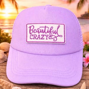 Puede incluir: Gorra de camionero lavanda con parte trasera de malla y visera curva. La parte delantera de la gorra presenta un parche rectangular bordado con las palabras "Beautiful Crazy" en escritura púrpura.