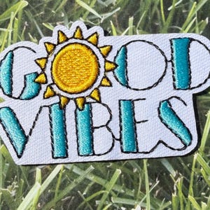 Puede incluir: Parche bordado con las palabras "GOOD VIBES" en letras turquesas con contorno negro. La letra "O" es un círculo, y el sol es amarillo con contornos negros. El parche está sobre un fondo blanco.