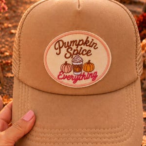 以下が含まれることがあります： Pumpkin Spice Everythingという文字、ホイップクリームをのせたコーヒーカップ、2つのカボチャのイラストが描かれた円形のパッチが付いた、タンカラーのトラッカーキャップ。前面は無地で、背面はメッシュ素材になっています。