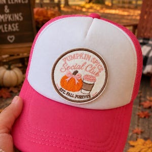 May include: A bright pink and white trucker hat featuring a circular embroidered patch on the front. The patch displays an orange pumpkin with a pink bow and a coffee cup, with the text PUMPKIN SPICE SOCIAL CLUB and EST. FALL FOREVER.