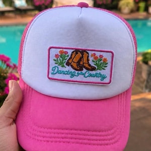 May include: A pink and white trucker hat with a patch that reads "Dancing in the Country." The patch features brown cowboy boots and orange flowers. The hat has a pink brim and a white mesh back.