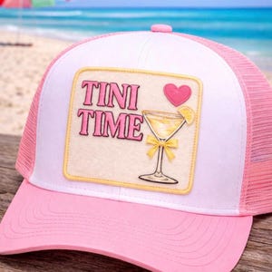 May include: Pink and white trucker hat with a pink brim and mesh back. The hat features a patch with the words "TINI TIME" above a cocktail illustration, a pink heart, and a lemon slice. The background shows a beach scene.