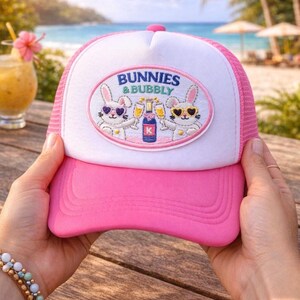 May include: A pink and white trucker hat with the text "Bunnies & Bubbly" on the front. The hat features an embroidered patch with two cartoon bunnies wearing sunglasses and holding champagne glasses. The hat is held up by a person's hands.