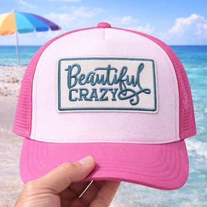 Peut inclure: Casquette de camionneur rose et blanche avec l'inscription "Beautiful Crazy" brodée sur un patch rectangulaire. La casquette a une visière rose et un dos en filet. L'arrière-plan montre une plage avec un parasol coloré.