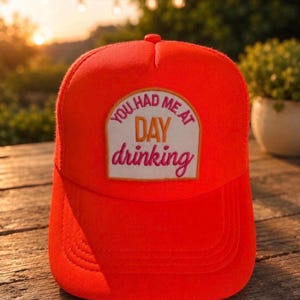 May include: Bright orange trucker hat with a curved brim. The hat features a rectangular patch with the text "YOU HAD ME AT DAY drinking" in white and pink lettering. The hat is sitting on a wooden surface.