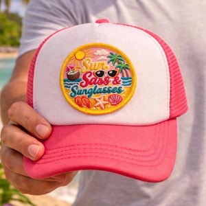 May include: A pink and white trucker hat with a circular patch featuring the text "Sun, Sass & Sunglasses" with a beach theme. The hat has a pink brim and mesh back, perfect for sunny days.