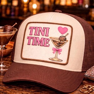 May include: A brown and cream trucker hat with a rectangular patch. The patch displays the words "TINI TIME" in pink, a pink heart, and a cocktail illustration. The hat has a curved peak and mesh back.