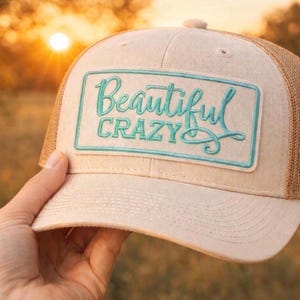 May include: A beige and brown trucker hat with the words "Beautiful Crazy" embroidered in teal on a rectangular patch. The hat has a curved brim and a mesh back.