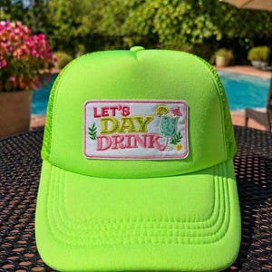 May include: Bright neon green trucker hat with a matching brim and mesh back. The hat features a rectangular patch with the words "LET'S DAY DRINK" and a cocktail illustration. The hat is designed for casual wear.
