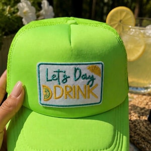 Könnte beinhalten: Leuchtend neongrüne Trucker-Cap mit gebogenem Schirm. Auf der Cap befindet sich ein rechteckiger Aufnäher mit dem Text "Let's Day DRINK" und einer Zitronengrafik. Ein leuchtendes Accessoire für Outdoor-Aktivitäten.