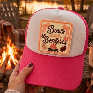 May include: A bright pink trucker hat with a white front panel featuring a square patch. The patch displays the text "Bows and Bonfires" in dark brown script, surrounded by embroidered illustrations of two peach bows, autumn leaves, and a campfire.