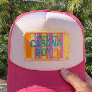 Puede incluir: Gorra de camionero rosa y blanca con la frase bordada "Where's my Cabana Boy". La gorra tiene una visera rosa y un panel frontal blanco. El texto es en turquesa y amarillo.