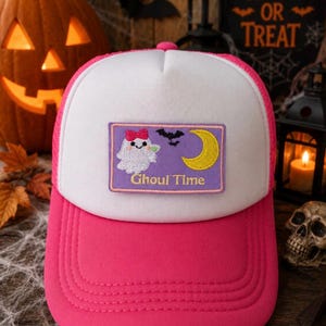 May include: A hot pink and white trucker hat featuring a rectangular patch on the front. The patch is purple with an embroidered white ghost wearing a pink bow, a black bat, a yellow crescent moon, and the text Ghoul Time in yellow lettering.