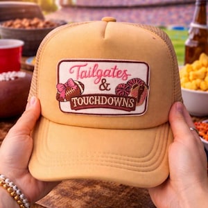 May include: A tan trucker hat with a mesh back and a curved brim. The front patch reads "Tailgates & Touchdowns" with football and donut graphics in pink and brown. The hat is held by hands, suggesting it's ready to wear.