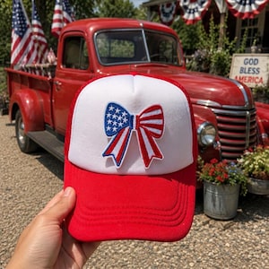 May include: A red and white trucker hat featuring a central patch shaped like a bow. The bow design incorporates the American flag pattern with blue and white stars on the left side and red and white stripes on the right side, finished with a red border.
