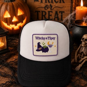 May include: A black and white trucker hat featuring a rectangular embroidered patch on the front panel. The patch displays the text Witchy & Tipsy in purple, accompanied by an embroidered purple witch hat, a cauldron, and a cocktail glass.