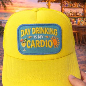 May include: Yellow trucker hat with a blue patch that reads "Day Drinking is my Cardio." The patch features a cocktail, lightning bolts, and a sun. The hat is held up in front of a beach bar at sunset.