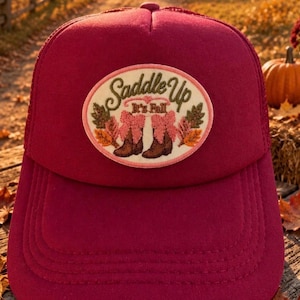 Puede incluir: Una gorra tipo trucker de color rojo oscuro con un parche ovalado central que presenta un diseño bordado de botas vaqueras marrones con lazos rosas, rodeadas de hojas de otoño y el texto "Saddle Up It's Fall" en cursiva verde oscuro.